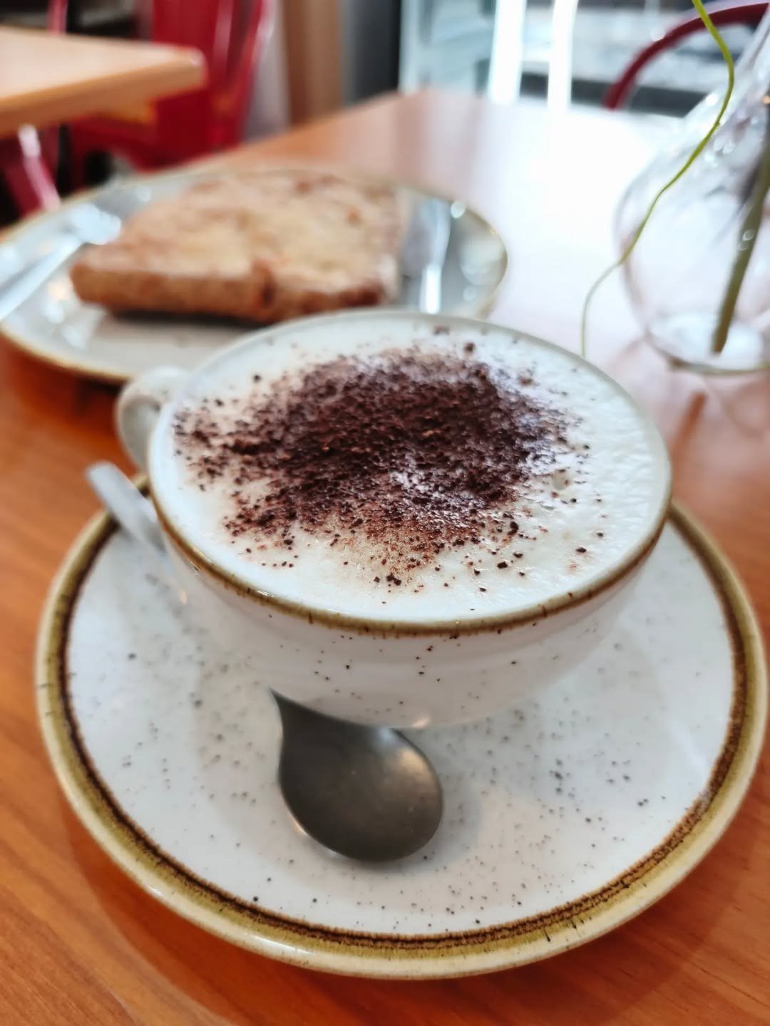 Cappuccino com cacau numa chávena branca, com uma torrada ao fundo
