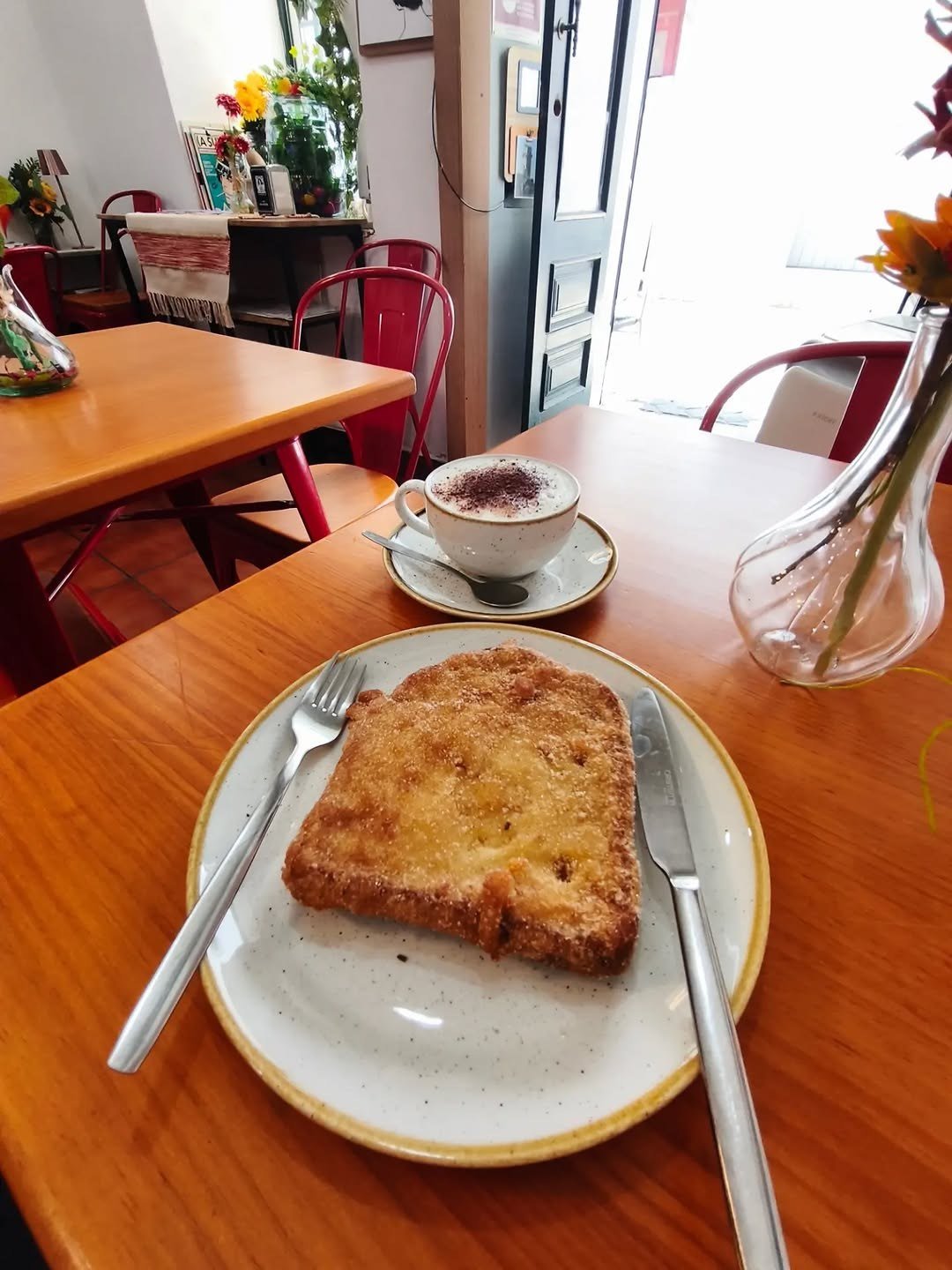 Mesa de madeira com torrada e cappuccino, cadeiras vermelhas e a porta aberta para a rua